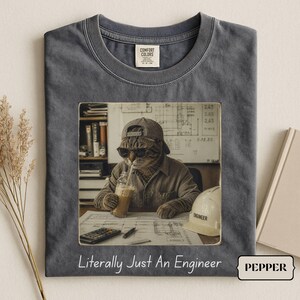 Peut inclure: Un t-shirt à col rond gris anthracite avec un imprimé représentant un chat portant une casquette et des lunettes de soleil, buvant dans un verre devant des plans et un casque de chantier. Le texte sous l'image indique, Literally Just An Engineer.