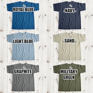 Puede incluir: Un collage de 12 camisetas de diferentes colores, incluyendo amarillo, azul real, azul marino, blanco, negro, azul claro, arena, rosa claro/coral, gris ceniza, grafito, verde militar y rojo. Cada camiseta tiene un color diferente y se muestra sobre un fondo de madera blanco.