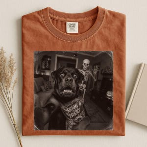 May include: A rust-colored crew neck t-shirt featuring a black and white graphic print of a dog holding a Trick or Treat bag with a skeleton standing in the background. The shirt has a Comfort Colors brand label on the inside of the collar.