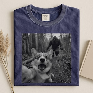 K&ouml;nnte beinhalten: Ein marineblaues T-Shirt mit Rundhalsausschnitt und einem schwarz-wei&szlig;en Grafikdruck eines Corgis, der ein Selfie in einem Wald macht, w&auml;hrend im Hintergrund eine Bigfoot-Figur zu sehen ist. Das Comfort Colors Etikett ist am Kragen.
