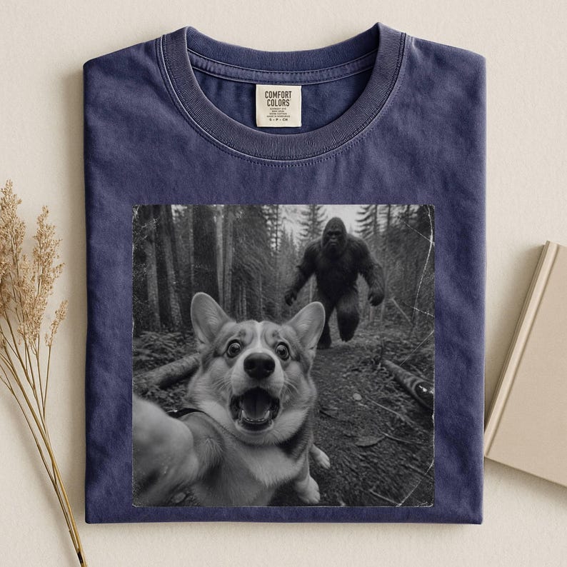 K&ouml;nnte beinhalten: Ein marineblaues T-Shirt mit Rundhalsausschnitt und einem schwarz-wei&szlig;en Grafikdruck eines Corgis, der ein Selfie in einem Wald macht, w&auml;hrend im Hintergrund eine Bigfoot-Figur zu sehen ist. Das Comfort Colors Etikett ist am Kragen.