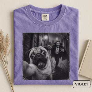Puede incluir: Una camiseta morada de cuello redondo con un estampado gráfico en blanco y negro de un pug tomándose una selfie en un callejón oscuro con una figura de vampiro al fondo. La camiseta tiene una etiqueta de la marca Comfort Colors en el cuello.