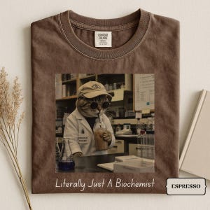 Könnte beinhalten: Ein braunes T-Shirt mit Rundhalsausschnitt und einer Grafik eines Katzen-Biochemikers im Laborkittel, mit Baseballkappe und runder Sonnenbrille, der ein Getränk hält. Unter der Grafik steht der Text, Literally Just A Biochemist.