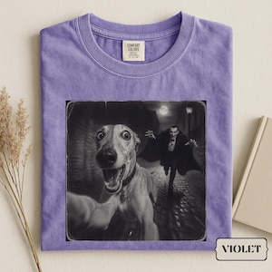 Può includere: Una t-shirt viola a girocollo con una stampa grafica che ritrae un cane che scatta un selfie in primo piano e una figura di vampiro che corre sullo sfondo di una strada acciottolata. La maglietta ha un'etichetta Comfort Colors sul colletto.