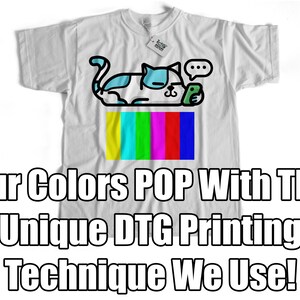 May include: White t-shirt with a cartoon cat design holding a phone. The cat is sitting on top of a colorful television screen with the text "Our Colors POP With The Unique DTG Printing Technique We Use!"