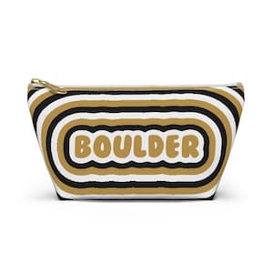 May include: A black and white striped makeup bag with gold accents. The bag has the word "BOULDER" written in a retro font.