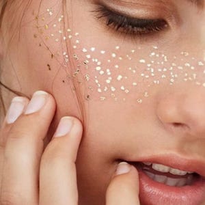 May include: A close-up view of a face featuring small, metallic gold flecks applied to the skin across the bridge of the nose and cheeks, mimicking the appearance of freckles. The gold particles are irregular in shape and size, creating a shimmering effect on the skin surface.