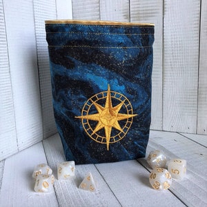 May include: A blue and gold fabric dice bag with a compass design. The bag is filled with white dice with gold numbers.