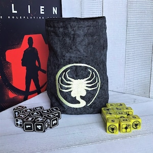 May include: A black fabric dice bag with an embroidered alien design. The bag is filled with black dice with white numbers. The bag is sitting on a white wooden surface.