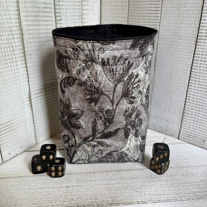 May include: A black and white floral patterned fabric dice bag with a black lining. Four black dice with white pips are sitting in front of the bag.