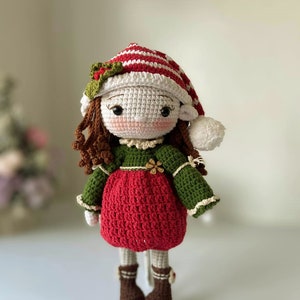 May include: A crocheted doll wearing a red and white striped hat with a green sweater and red dress. The doll has brown hair and brown boots.
