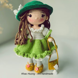 May include: A crocheted doll wearing a green hat, a white dress with a green skirt, and brown hair. The doll is holding a small brown purse with a white flower.