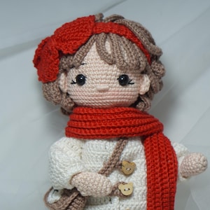 以下が含まれることがあります： 赤いスカーフ、帽子、ヘアバンドをつけた手作りのかぎ針編み人形。人形は木製のボタンが付いた白いコートと小さな茶色のバッグを持っています。人形の髪は茶色で、肌は明るいピーチ色です。