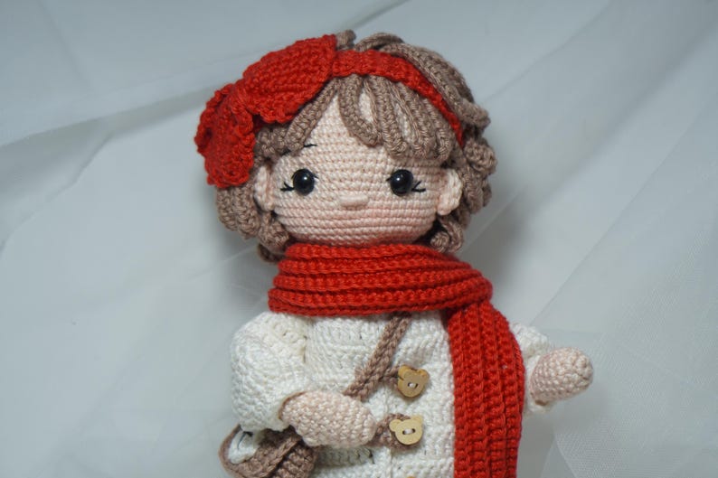 以下が含まれることがあります： 赤いスカーフ、帽子、ヘアバンドをつけた手作りのかぎ針編み人形。人形は木製のボタンが付いた白いコートと小さな茶色のバッグを持っています。人形の髪は茶色で、肌は明るいピーチ色です。
