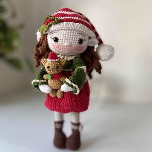 May include: A crocheted elf doll wearing a red and white striped hat with a green holly sprig. The doll is holding a crocheted brown teddy bear wearing a red and white hat.
