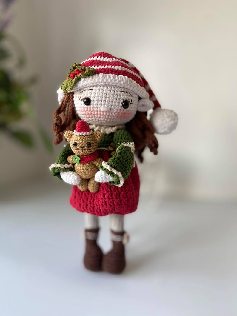 May include: A crocheted elf doll wearing a red and white striped hat with a green holly sprig. The doll is holding a crocheted brown teddy bear wearing a red and white hat.
