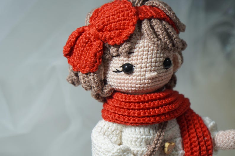 以下が含まれることがあります： 赤いリボン、スカーフ、ヘアバンドが付いたかぎ針編みの人形。人形は白いドレスと茶色の髪をしています。人形の目は黒く、スカーフは首に巻かれています。人形は手作りです。