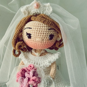 Puede incluir: Una mu&ntilde;eca de crochet vestida con un vestido de novia blanco con velo y sosteniendo un ramo de flores rosas. La mu&ntilde;eca tiene el pelo casta&ntilde;o, la cara rosa y los ojos negros.