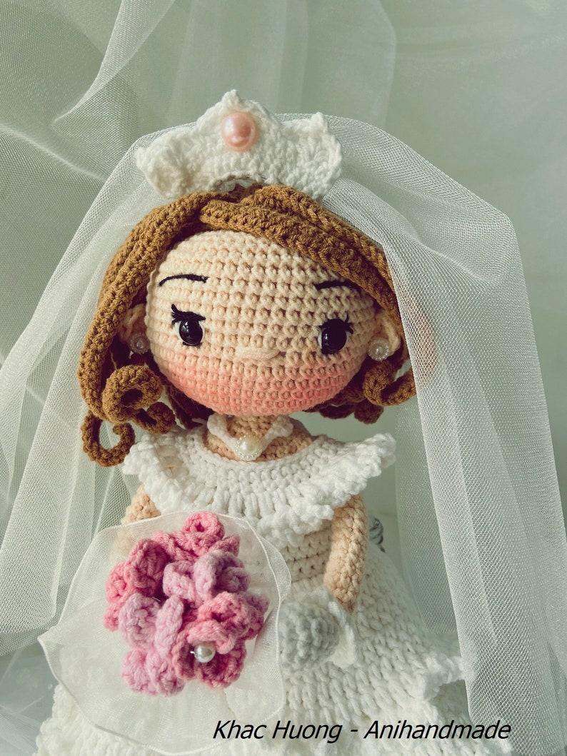 Puede incluir: Una mu&ntilde;eca de crochet vestida con un vestido de novia blanco con velo y sosteniendo un ramo de flores rosas. La mu&ntilde;eca tiene el pelo casta&ntilde;o, la cara rosa y los ojos negros.