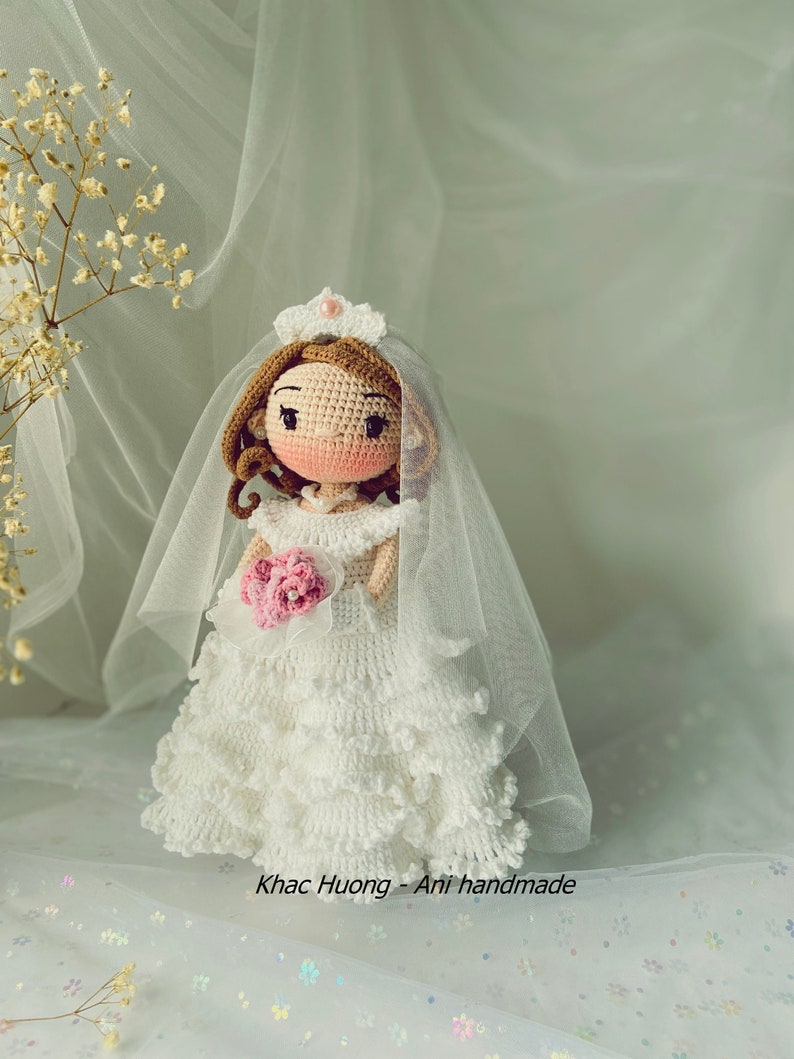 Puede incluir: Una mu&ntilde;eca de novia de crochet que lleva un vestido blanco con velo y sostiene un ramo de flores rosas. La mu&ntilde;eca tiene el pelo casta&ntilde;o y la cara rosa. La mu&ntilde;eca est&aacute; de pie sobre un fondo blanco con un patr&oacute;n floral.
