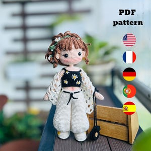 Könnte beinhalten: Gehäkelte Puppe mit einem schwarzen Oberteil mit weißen Gänseblümchen-Akzenten, einer weißen Hose und einer weißen gehäkelten Jacke. Die Puppe hat braune Haare und hält eine schwarze Handtasche. Das Bild enthält den Text "PDF pattern" und Flaggen.