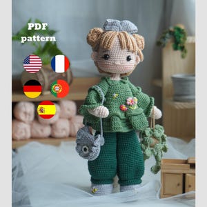 May include: A crochet doll with light brown hair in buns, wearing a green ruffled top with flower embroidery, dark green trousers, and grey shoes. The doll carries a grey bag and a small potted plant. Text reads PDF pattern with flags.