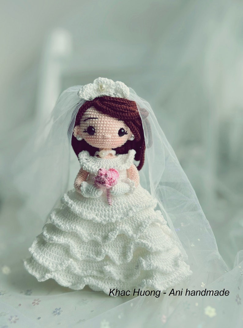 Puede incluir: Una mu&ntilde;eca de novia de crochet con un vestido blanco, velo y un ramo de flores rosas. La mu&ntilde;eca tiene el pelo casta&ntilde;o, ojos marrones y un collar de perlas. La mu&ntilde;eca est&aacute; de pie sobre una superficie blanca con un patr&oacute;n floral.