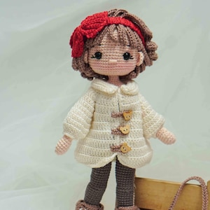 以下が含まれることがあります： 手作りのかぎ針編み人形。茶色の髪に赤いリボン。クリーム色のコート、クマの形をしたボタン、茶色のレギンス、茶色の靴を着用。クマの顔の小さなハンドバッグも含まれています。「Khac Huong - Ani handmade」の文字が見えます。