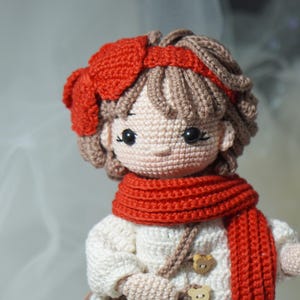 以下が含まれることがあります： 赤いリボン、スカーフ、ヘアバンドが付いた手作りの編み人形。人形は茶色の髪で、木製のボタンが付いた白いドレスを着ています。人形の顔は黒い目と薄いピンクの肌色で詳細に描かれています。
