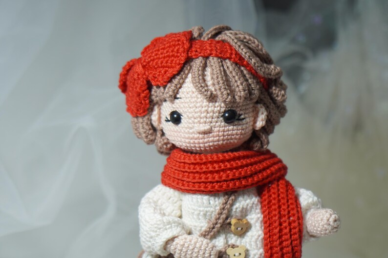 以下が含まれることがあります： 赤いリボン、スカーフ、ヘアバンドが付いた手作りの編み人形。人形は茶色の髪で、木製のボタンが付いた白いドレスを着ています。人形の顔は黒い目と薄いピンクの肌色で詳細に描かれています。