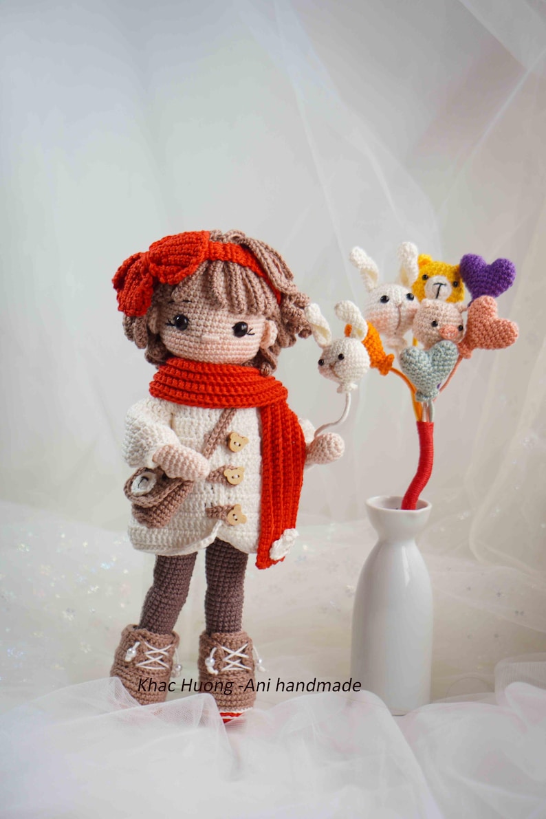 以下が含まれることがあります： 白いコート、赤いスカーフ、ヘアバンドを身に着けた手作りのかぎ針編み人形。人形は小さなバッグとかぎ針編みの動物とハートのブーケを持っています。「Khac Huong - Ani handmade」の文字が見えます。