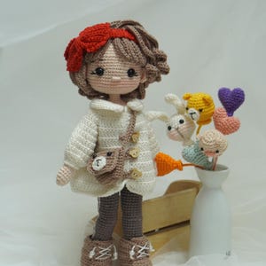 以下が含まれることがあります： 手作りのかぎ針編み人形。茶色の髪に赤いリボン。白いコート、茶色のズボン、ブーツを着用。クマのデザインの小さな茶色のバッグが肩からぶら下がっています。花瓶にはかぎ針編みの動物とハートの装飾が飾られています。