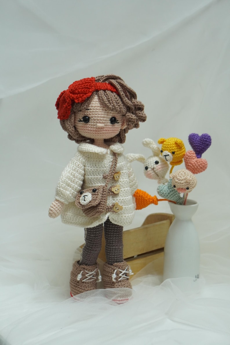 以下が含まれることがあります： 手作りのかぎ針編み人形。茶色の髪に赤いリボン。白いコート、茶色のズボン、ブーツを着用。クマのデザインの小さな茶色のバッグが肩からぶら下がっています。花瓶にはかぎ針編みの動物とハートの装飾が飾られています。
