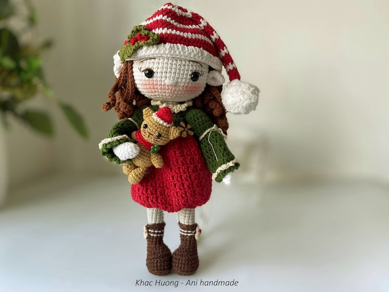 May include: A crocheted elf doll wearing a red and white striped hat with a white pom-pom. The doll is holding a crocheted brown teddy bear. The doll is wearing a red dress with a green crocheted sweater and brown boots.