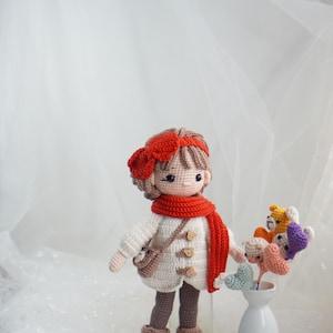 以下が含まれることがあります： 白いコート、赤いスカーフ、茶色のブーツを履いた手作りのかぎ針編みの人形。人形は赤いリボンと小さなバッグを持っています。花瓶には、さまざまな色の編み物の花がいくつか入っています。