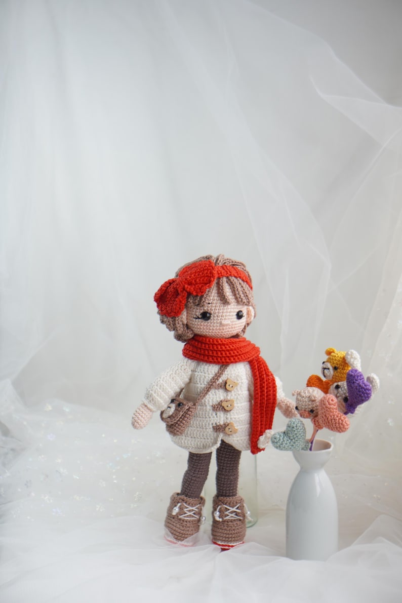 以下が含まれることがあります： 白いコート、赤いスカーフ、茶色のブーツを履いた手作りのかぎ針編みの人形。人形は赤いリボンと小さなバッグを持っています。花瓶には、さまざまな色の編み物の花がいくつか入っています。