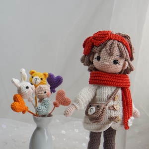 以下が含まれることがあります： 赤いリボン、スカーフ、ブーツを身に着けた手作りのかぎ針編みの人形。かぎ針編みの花瓶の隣にあります。人形は白いセーターと茶色のハンドバッグを着ています。花にはウサギ、クマ、豚、ハートが含まれています。
