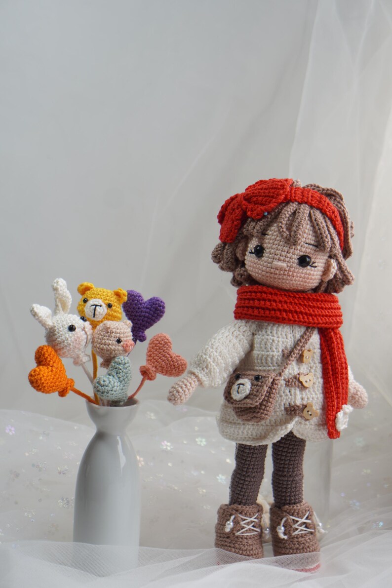 以下が含まれることがあります： 赤いリボン、スカーフ、ブーツを身に着けた手作りのかぎ針編みの人形。かぎ針編みの花瓶の隣にあります。人形は白いセーターと茶色のハンドバッグを着ています。花にはウサギ、クマ、豚、ハートが含まれています。