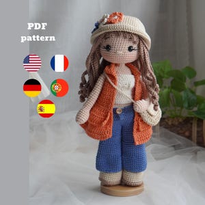 Könnte beinhalten: Eine Häkelpuppe mit langem braunem Haar, einem beigen Hut mit orangefarbenen und blauen Blumen, einem weißen Oberteil, einer orangefarbenen Weste, blauer Hose und beigen Schuhen. Die Puppe trägt eine kleine beige Tasche. Text PDF pattern und fünf Flaggen.