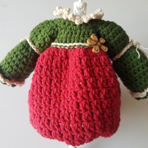 May include: A crocheted doll dress with long sleeves, a red bodice, a green top, and a white crocheted collar. The dress has a wooden flower embellishment.