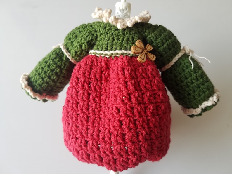 May include: A crocheted doll dress with long sleeves, a red bodice, a green top, and a white crocheted collar. The dress has a wooden flower embellishment.