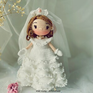 Puede incluir: Una mu&ntilde;eca de crochet vestida con un vestido de novia blanco con velo y corona. La mu&ntilde;eca tiene el pelo casta&ntilde;o, la cara rosa y los ojos negros. La mu&ntilde;eca lleva un ramo de flores rosas.