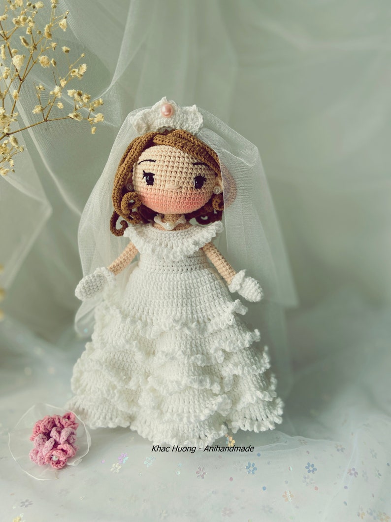 Puede incluir: Una mu&ntilde;eca de crochet vestida con un vestido de novia blanco con velo y corona. La mu&ntilde;eca tiene el pelo casta&ntilde;o, la cara rosa y los ojos negros. La mu&ntilde;eca lleva un ramo de flores rosas.