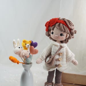 以下が含まれることがあります： 茶色の髪と赤いヘアバンドをした手作りのかぎ針編みの人形。白いセーターと茶色のブーツを着用。クマのデザインの小さなバッグが肩からぶら下がっています。花瓶にはかぎ針編みの花と動物が入っています。