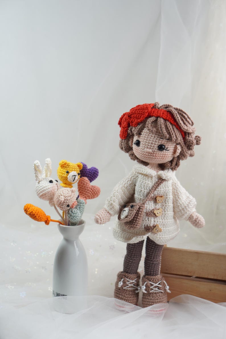 以下が含まれることがあります： 茶色の髪と赤いヘアバンドをした手作りのかぎ針編みの人形。白いセーターと茶色のブーツを着用。クマのデザインの小さなバッグが肩からぶら下がっています。花瓶にはかぎ針編みの花と動物が入っています。