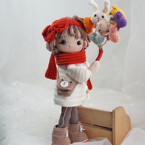 以下が含まれることがあります： 赤いスカーフとヘアバンドを身につけ、カラフルな編みぐるみの動物のブーケを持っている手作りの編みぐるみ人形。人形は白いドレス、茶色のブーツ、クマの小さな財布を着ています。木製の箱の前に立っています。