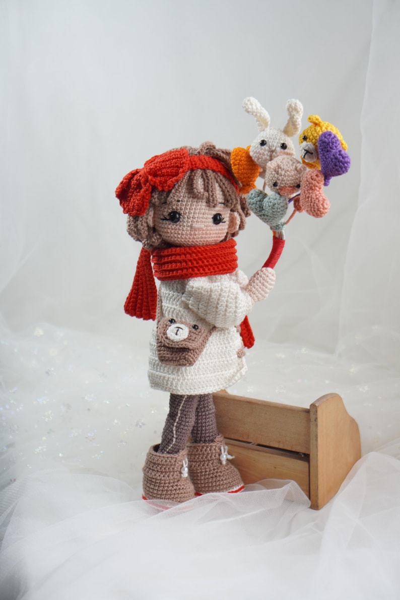 以下が含まれることがあります： 赤いスカーフとヘアバンドを身につけ、カラフルな編みぐるみの動物のブーケを持っている手作りの編みぐるみ人形。人形は白いドレス、茶色のブーツ、クマの小さな財布を着ています。木製の箱の前に立っています。