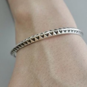 May include: A silver bracelet featuring a repeating triangular pattern. The bracelet is displayed on a wrist, highlighting the detailed design and metallic finish. The triangles are evenly spaced around the band.