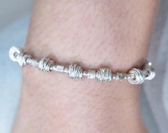 May include: A silver bracelet with a unique design. The bracelet features a series of textured, spherical beads and square links, connected by a delicate chain. The bracelet is worn on a wrist, showcasing its intricate details and craftsmanship.