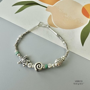 May include: Silver bracelet with a starfish, spiral, and fish charm. The bracelet has turquoise beads and decorative elements. The image includes the text "V6B019 10.6 g 6.5 ". The bracelet is approximately 16.5 cm in length.